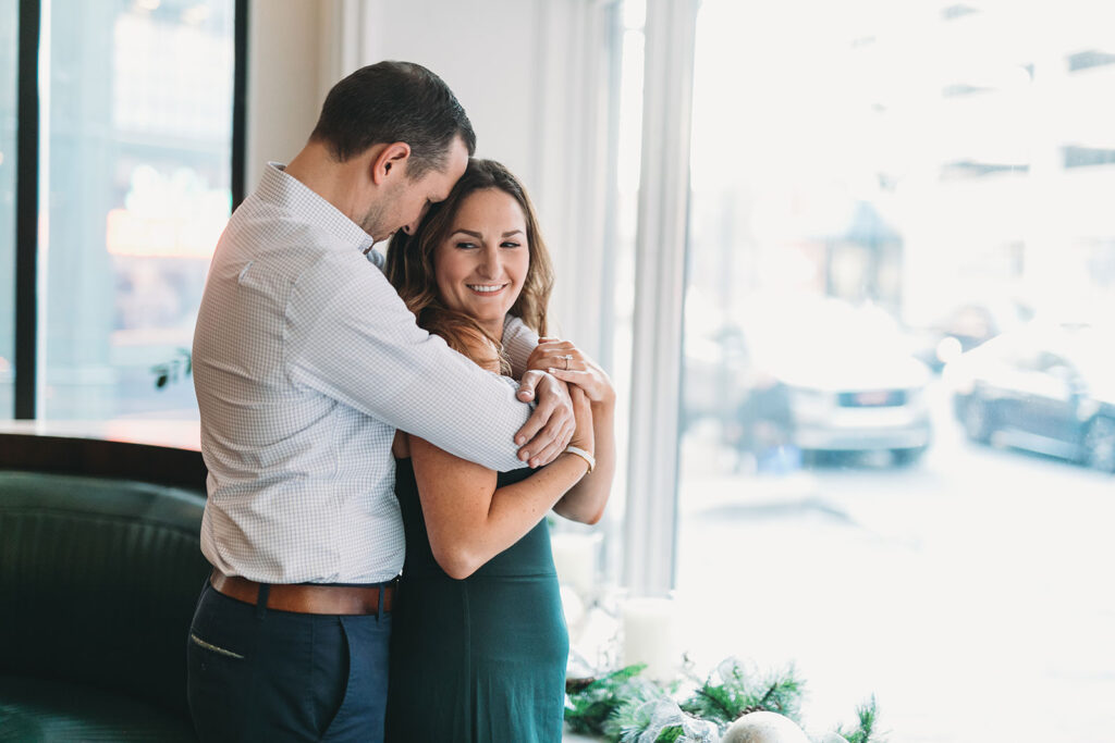 man hugs woman by window facing street with cars during their le meridien engagement photo session